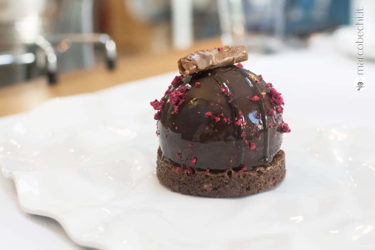 Sfera al cioccolato su pan di spagna al cacao, all'interno cremoso al caffè, mousse alla nocciola e lampone disidratato.